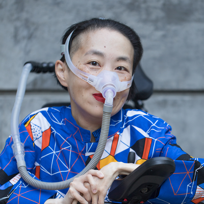Photo of an Asian American woman in a power chair. She is wearing a blue shirt with a geometric pattern with orange, 黑色的, 白色, and yellow lines and cubes. She is wearing a mask over her nose attached to a gray tube and bright red lip color. She is smiling at the camera. Photo credit: Eddie Hernandez Photography