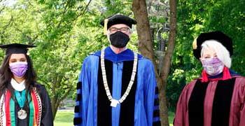 President Glotzbach, Jennifer Mueller, and Jinan Al-Busaidi stand for a photo with graduation outfits and masks
